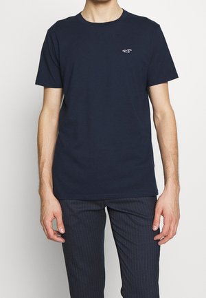 Mann trägt ein marineblaues T-Shirt mit kurzen Ärmeln, einem kleinen weißen Logo auf der Brust und dunkelgestreifte Hosen.