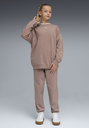 Junges Mädchen mit geflochtenem Haar, das einen lockeren taupefarbenen Pullover und passende Hose trägt, steht mit einer Hand, die ihre Flechte berührt.