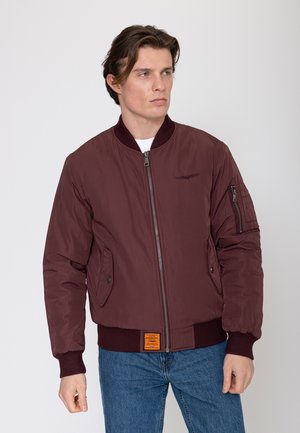 Homme portant une veste bomber bordeaux avec une fermeture éclair et des poches à boutons, debout devant un fond clair uni, regardant vers la droite.