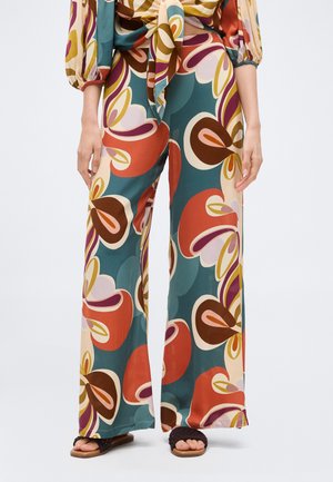 Pantalon taille haute à jambes larges avec grand imprimé floral abstrait en bleu sarcelle, orange, marron et rose, porté avec des sandales noires à bouts ouverts.