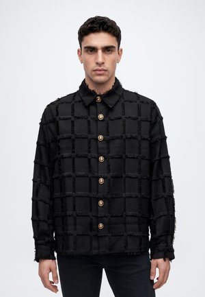 Un homme porte une veste noire à franges, à motif quadrillé et ornée de boutons dorés, sur un fond clair et uni.