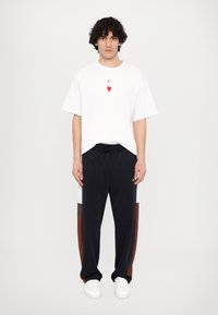 Tricou scurt alb cu mânecă, având un grafic în formă de inimă roșie, asortat cu pantaloni de trening bleumarin, care au panouri laterale colorate în nuanțe de maro și alb. Adidași albi.