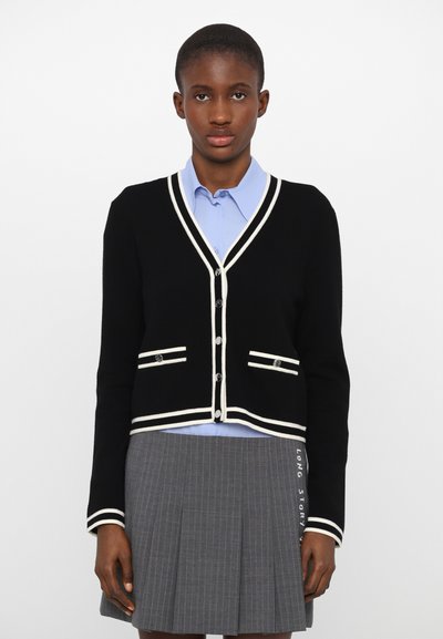 Schwarze Strickjacke mit weißem Rand, V-Ausschnitt und zwei Taschenakzenten. Über einem hellblauen Hemd getragen, kombiniert mit einem grauen Faltenrock.