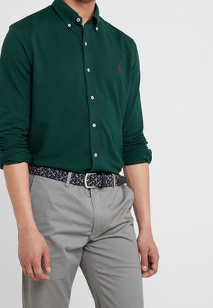 Homme portant une chemise boutonnée vert foncé avec les manches retroussées, un pantalon gris clair et une ceinture tissée noire et grise, debout devant un fond uni.