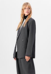Frau mit langen blonden Haaren, die einen dunkelgrauen oversized Anzug und ein schwarz-weißes gepunktetes Shirt trägt, schaut seitlich vor einem weißen Hintergrund.