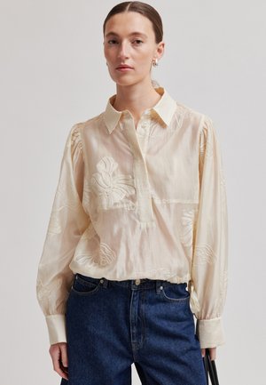 Woman wearing sheer cream blouse with floral embroidery and long sleeves, paired with high-waisted dark blue jeans, holding black bag.