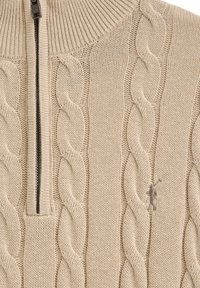 Beige kabelstickad tröja med hög krage och halvdragkedja. Har ett strukturerat mönster och en diskret logo broderad i mörkare beige.