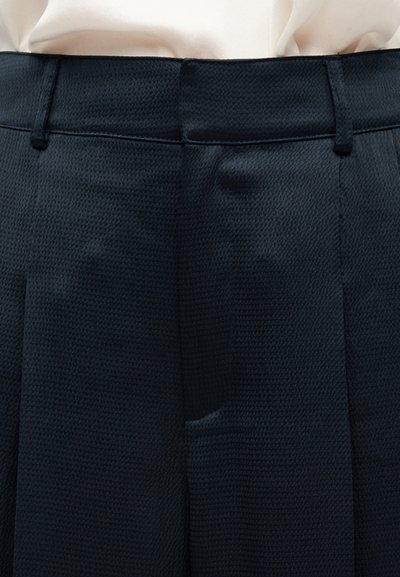 Κοντινό πλάνο σκούρων υφασμάτινων παντελονιών με θηλιές για ζώνη, φορεμένα με ανοιχτόχρωμο πουκάμισο μέσα από το παντελόνι.