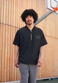 Sort kortærmet polo skjorte med lynlås, der har en frontlomme med et logo og en afslappet pasform, parret med grå bukser.