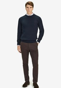 Tummansininen palmikkoneule, jossa on pyöreä kaula-aukko, käytetty valkoisen kauluspuseron päällä, yhdistetty ruskeisiin housuihin ja tummiin mokkakenkiin.