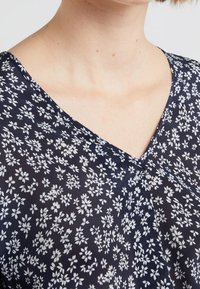 Laivansininen kukkasaalinen, jossa on V-pääntie valkoisilla kukkakuoseilla, valmistettu kevyestä materiaalista, jolla on sileä pinta.