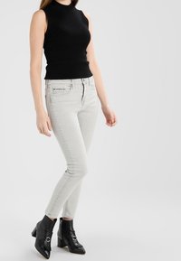 Svart ribbad ärmlös topp, ljusgrå figurnära jeans och svarta klackade ankelstövlar med en spänndetalj. Enkelt bakgrund.