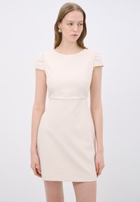 Hihaton, istuvan muotoisen mekon vaaleanbeige kangas. Sileä pinta, pyöreä pääntie, pussit olkapäillä ja suora leikkaus helmaan.