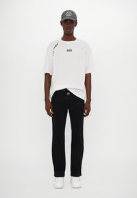 T-shirt blanc surdimensionné avec des accents sur les côtés, pantalon noir avec coutures blanches et casquette grise. Le modèle porte des baskets blanches. Design simple et moderne.