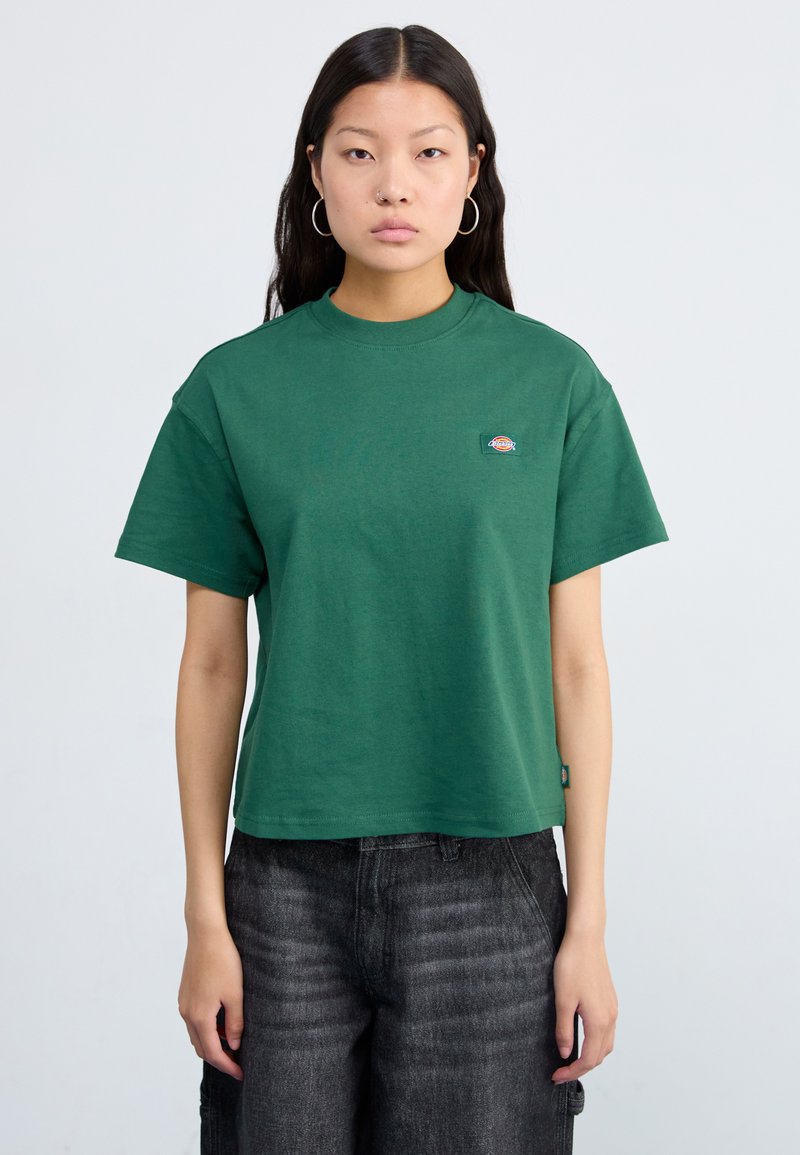 T-shirt in cotone verde scuro con collo rotondo, maniche corte e un piccolo logo sulla parte sinistra del petto. Indossato con pantaloni grigi scuri.