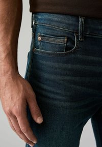 Kék farmernadrág keskeny szabással, réz szegecsekkel, elülső zsebbel és enyhe kopással. Kéz nyugodtan a combon.