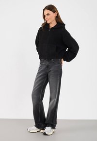 Felpa con cappuccio nera in pile con chiusura a zip, vestibilità oversize e tasche frontali. Abbinata a jeans grigi a gamba larga e sneaker sportive grigie.