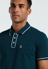 Polo de couleur sarcelle en tissu lisse, avec un col classique à bord blanc, trois boutons, une poche poitrine et un logo de pingouin.