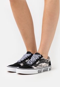 Benen bär svart och vit Vans-sneakers med mönstrade snören och grafisk text på sidorna och sula kanten.