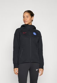 Svart zip-up hettegenser med en tettsittende design, med en rød Nike-logo og et blått Paris Saint-Germain-emblemm. Myk tekstur, to sidelommer.