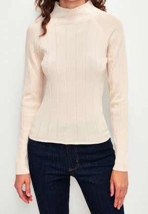 Femme portant un pull à col roulé côtelé crème ajusté à manches longues et un jean bleu foncé devant un fond uni.