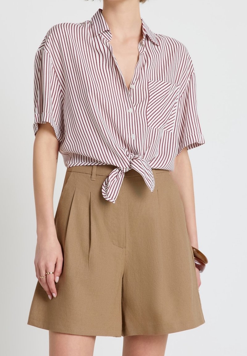 Femme portant une chemise à manches courtes blanche et rouge rayée nouée à la taille, associée à un short marron taille haute.