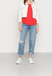 Giacca in denim bianco, top rosso con volant, jeans azzurri chiari strappati con dettagli di spruzzi e sneakers bianche. Maniche arrotolate sui jeans.