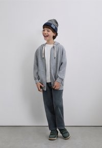 Grijze hoodie met ritssluiting en crèmekleurige trekkoorden, wit T-shirt, grijze corduroy broek en groene schoenen. Draagt een grijze en marineblauwe muts.