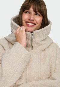 Beež fleece mantli kõrge kaelusega, lukuga sulgemine ja soonikkoes detailid. Pehme tekstuur ja ülekaaluline lõige.