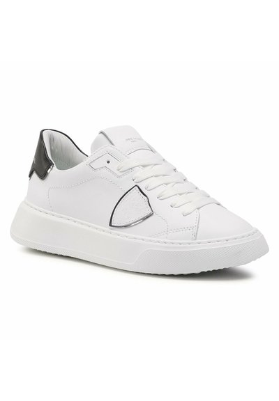 Sneaker bianca in pelle con finitura liscia, dettaglio del tallone nero, punta tonda e suola in gomma testurizzata. Presenta lacci bianchi piatti e un logo.