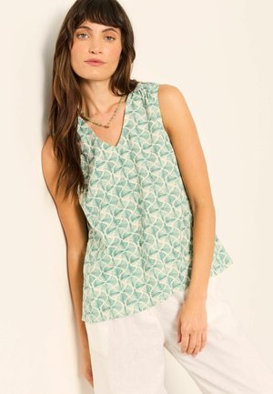 Young woman with long brown hair wearing a sleeveless green patterned top and white pants, standing against a light background.