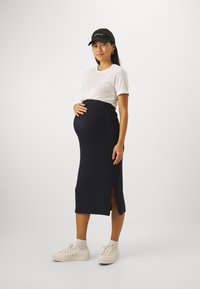 Jupe de maternité noire, côtelée, avec une fente sur le côté, associée à un t-shirt blanc et des baskets beiges. Le mannequin porte une casquette noire et des boucles d'oreilles texturées.