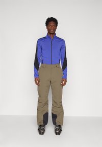 Μπλέ zip-up μπουφάν με μαύρες λεπτομέρειες, συνδυασμένο με καφέ παντελόνια που έχουν μαύρα κάτω μέρη. Το μοντέλο φορά μαύρες μπότες σκι.