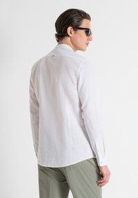 Camicia bianca in lino con maniche lunghe, polsini abbottonati, orlo arrotondato e un singolo dettaglio sul retro. Indossata con pantaloni verde chiaro.