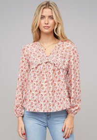 Blusa floreale con maniche lunghe, scollo a V e dettagli arricciati. Tessuto rosa chiaro decorato con piccoli fiori rossi e blu. Texture morbida.