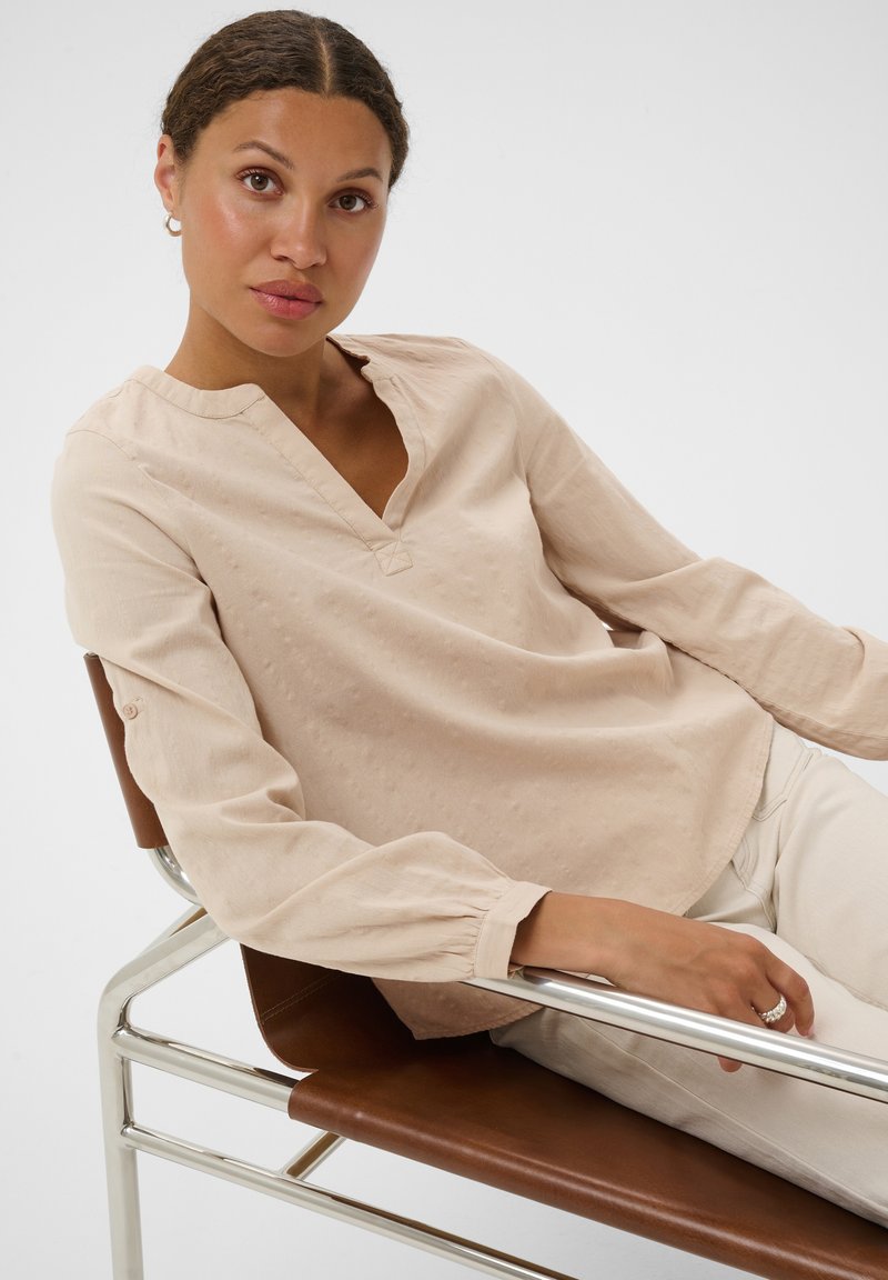 Femme aux cheveux tressés portant un chemisier beige à manches longues et un pantalon clair, assise de façon détendue sur une chaise en cuir marron et métal.