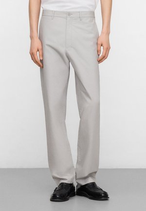 Mann trägt hellgraue, gerade geschnittene Hosen und schwarze Lederschuhe, steht vor einem schlichten hellen Hintergrund.