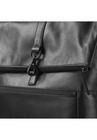 Sac en cuir noir avec une texture lisse, une fermeture éclair centrale et une bandoulière avec un accent matériel arrondi. Des coutures sont visibles.