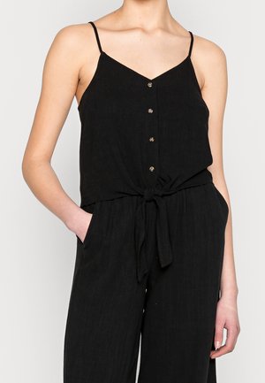 Kobieta w czarnym bezrękawnym kombinezonie z cienkimi ramiączkami, guzikami z przodu, przewiązanym paskiem w talii oraz kieszeniami po obu stronach.