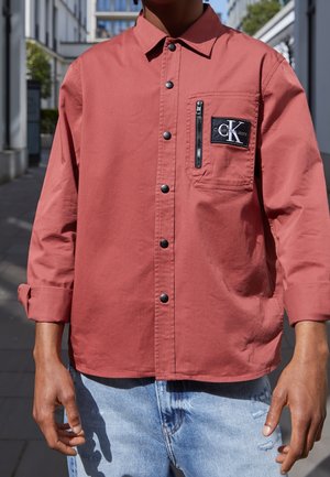 Person iført en langærmet, knappet, støvet rosa skjorte med lynlåslomme på brystet og Calvin Klein-logo, kombineret med lyseblå jeans.