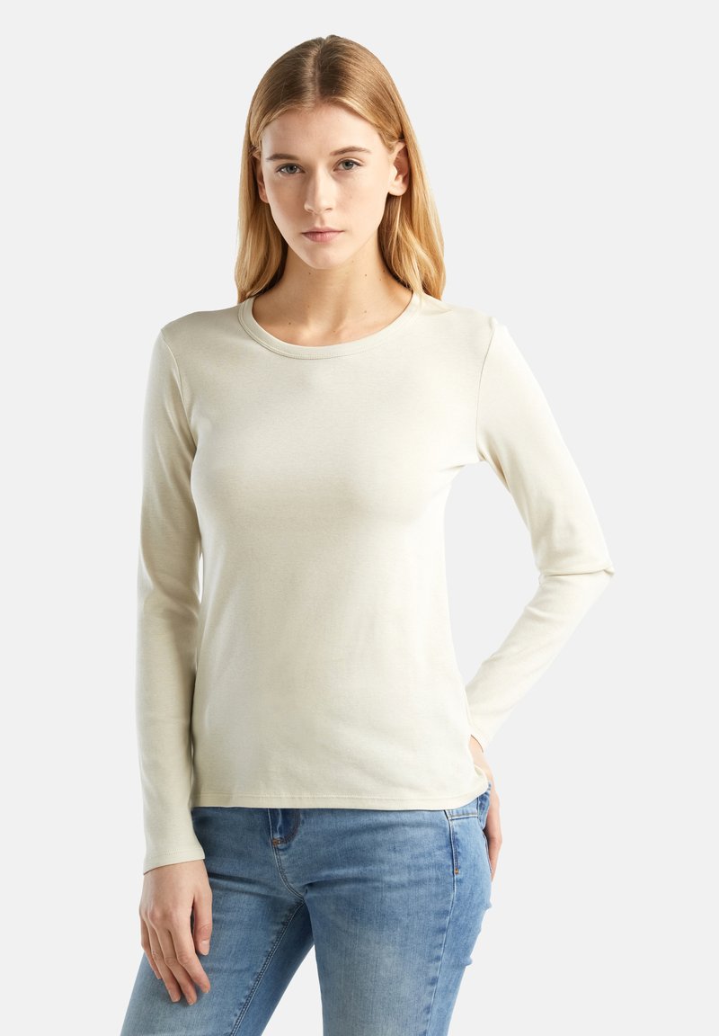 Donna con capelli biondi, che indossa una maglietta a maniche lunghe color crema e jeans blu, in piedi con una mano sulla vita contro uno sfondo neutro.