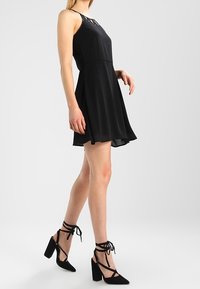 Woman wearing a sleeveless black dress with sheer hem and black high-heeled ankle-tie shoes, standing in a white studio.