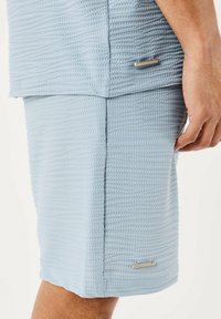 Los pantalones cortos de tela texturizada en azul claro presentan un corte holgado, costuras cuidadas y detalles metálicos a los lados, lo que realza el interés visual.