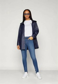 Mulher com cabelo trançado veste um casaco de chuva azul marinho, uma t-shirt branca, jeans azuis, ténis brancos e óculos de sol, de pé com uma mão no bolso.