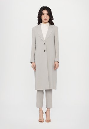 Vaaleanharmaa muotopuku, jossa on kaulusklaffi, kaksi nappia ja sivutaskut, yhdistettynä slim-fit-housuihin ja remmikenkiin.
