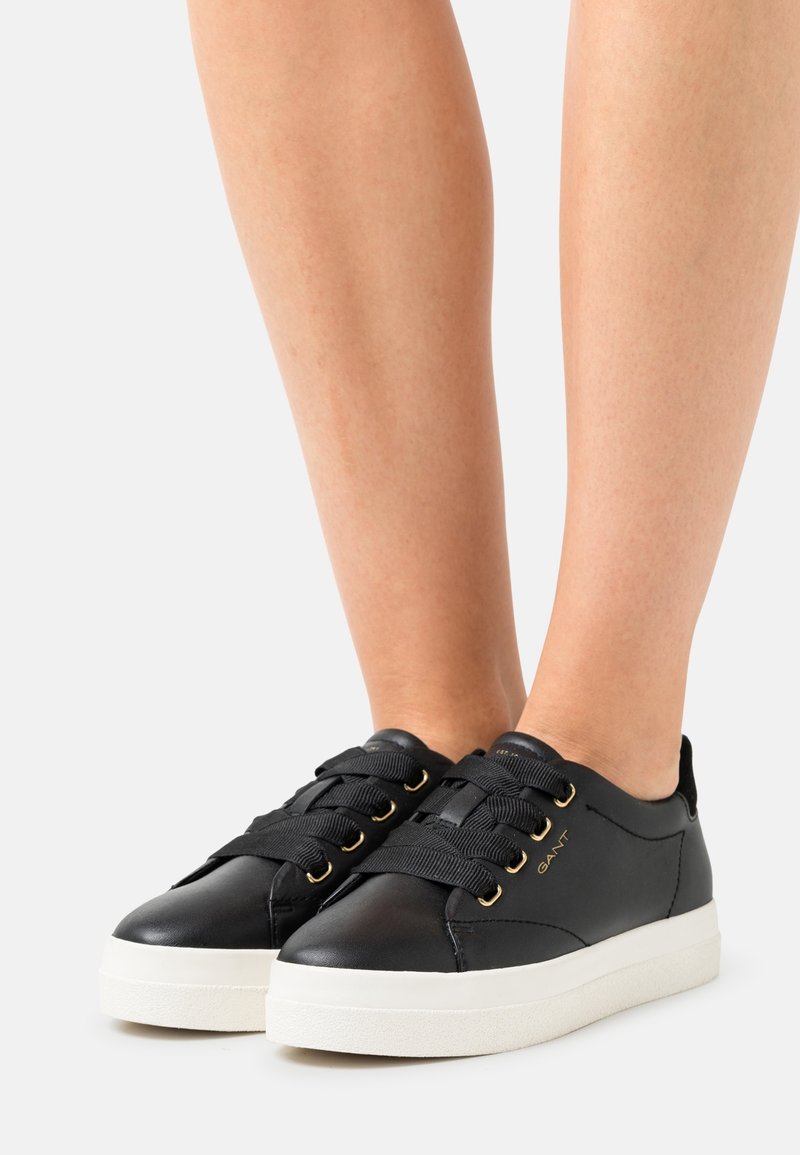 Baskets en cuir noir avec de larges lacets noirs, œillets dorés et semelles blanches portées pieds nus sur un fond uni.