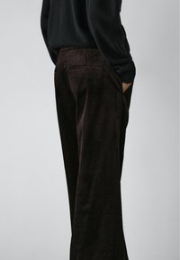 Person iført mørkebrune fløjlsbukser med vide ben og en sort sweater, stående med hænderne i lommerne mod en ensfarvet grå baggrund.