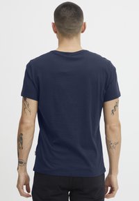 Mørkeblå bomull t-skjorte med rund hals, korte ermer, og en etikett synlig på venstre side, med detaljerte tatoveringer på begge armene.