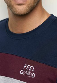 Primer plano de la parte inferior de la cara y el cuello de un hombre que lleva una camisa en tonos azul marino, burdeos y gris, con la frase "SIENTE BIEN" y un gráfico de bicicleta bordado en el pecho.