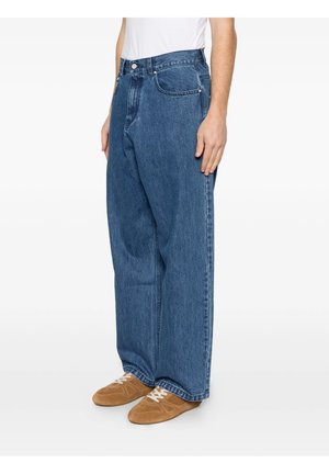 Persona che indossa jeans blu larghi con tasche visibili e sneakers in pelle scamosciata marrone con lacci bianchi, in piedi di lato.